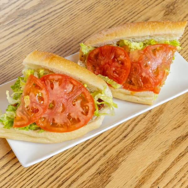 Two fresh sandwiches with sliced tomatoes and lettuce on a white plate, on a wooden table.